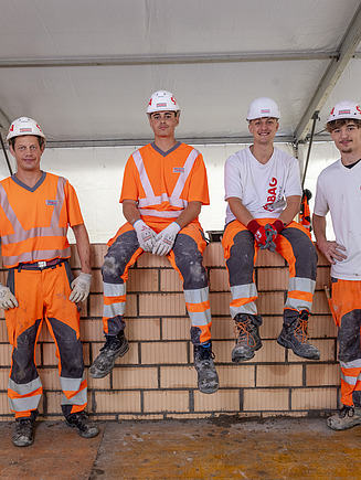 Drei STRABAG Lernende und ein Polier posieren stolz vor einer gemauerten Lochsteinwand in einem grossen Zelt. Zwei sitzen auf der Wand, zwei stehen daneben. Sie tragen orange Schutzkleidung und weisse STRABAG-Shirts.