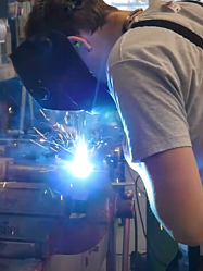 Junger STRABAG Lernender (Maschinist) beim Schweissen mit einer Schweisserbrille vor dem Gesicht. Im Hintergrund sieht man die Werkstatt.