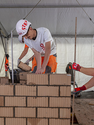 Zwei STRABAG Lernende beim Mauern ein Ziegelwand aus Lochsteinen. Einer setzt einen Ziegelstein, während der andere mit einem Gliedermassstab die Höhe überprüft. Die Arbeit findet in einem grossen Zelt statt.