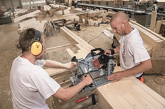 Lehrlinge stehen sich gegenüber und schneiden mit einer Spezialsäge mehrere Holzbalken auf Länge, beide mit Gehörschutz, im Hintergrund die Holzwerkstatt.
