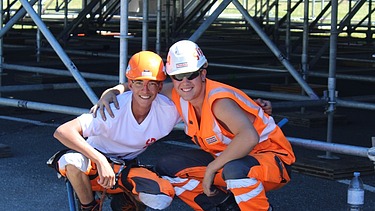 Zwei Lernende in Arbeitskleidung mit Helmen lächeln in die Kamera. Hinter ihnen ist ein Baugerüst aufgebaut.