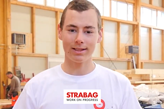 Junger STRABAG Lernender (Zimmermann) schaut lächeln in die Kamera in einem weissen Shirt. Im Hintergrund kann man die Holzbau-Werkstatt sehen.