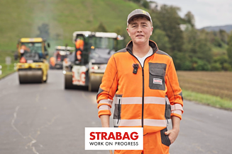 STRABAG Lernender mit Blick zur Kamera. Er trägt leuchtend-orange Arbeitskleidung mit STRABAG Logo. Im Hintergrund sind Bodenwalzen auf frisch asphaltierter Strasse im Einsatz.