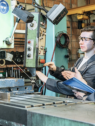 STRABAG Lernender bei der Arbeit, während der Praxisbildner kontrolliert.  Beide tragen graue Schutzkleidung an einer Fräsmaschine in der Werkstatt. Der Lernende bedient die Steuerung, der andere gibt Anweisungen und hält Unterlagen in der Hand. 