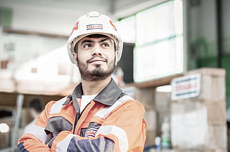 Ein junger STRABAG Lernender in orangener Sicherheitsjacke und Helm steht selbstbewusst mit verschränkten Armen in einer Werkhalle. Im Hintergrund sind Baumaterialien und STRABAG-Schilder zu erkennen.