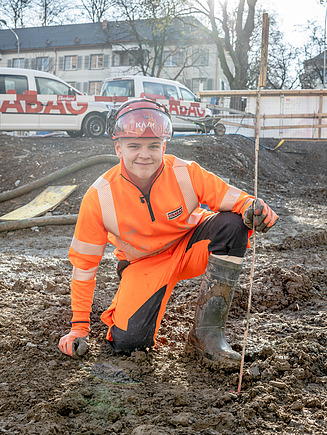 Junger STRABAG-Lernender in oranger Schutzkleidung kniet mit Gummistiefeln im Matsch und mit einem Massstab in der Hand – im Hintergrund STRABAG-Fahrzeuge und ein Wohnquartier.