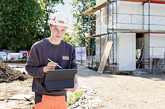 Eine Lehre auf der Baustelle - STRABAG deine Zukunft Ein junger STRABAG Lernender mit Helm und Arbeitskleidung steht auf einer Baustelle und arbeitet mit einem Tablet und einem digitalen Stift. Im Hintergrund ist ein eingerüstetes Gebäude sowie Baustellenmaterial zu sehen.