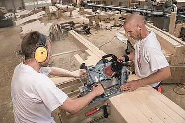 Lehre Zimmermann:Zimmerin Lehrlinge stehen sich gegenüber und schneiden mit einer Spezialsäge mehrere Holzbalken auf Länge, beide mit Gehörschutz, im Hintergrund die Holzwerkstatt.