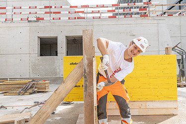 Lehre Maurer:in Junger STRABAG Lernender (Maurer) auf einer Baustelle. Er lächelt in die Kamera und hält einen Hammer und Nagel in der Hand. Mit weissem Helm und Shirt mit STRABAG Logo. Im Hintergrund sieht man eine Hochbaubaustelle