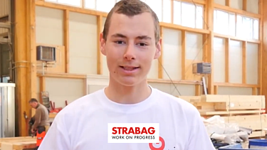 Junger STRABAG Lernender (Zimmermann) schaut lächeln in die Kamera in einem weissen Shirt. Im Hintergrund kann man die Holzbau-Werkstatt sehen.