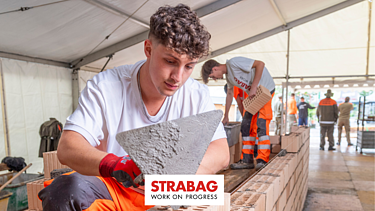 Ein STRABAG Lernender beim Mauern. Er trägt ein weisses STRABAG-Shirt und verarbeitet frischen Mörtel mit einer Kelle. Im Hintergrund sind weitere Lernende zu sehen.