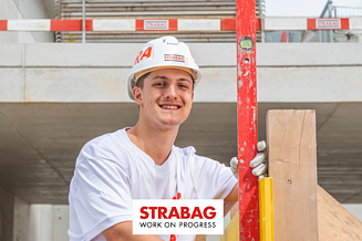Junger STRABAG Lernender (Maurer) auf einer Baustelle. Er lächelt in die Kamera und hält eine rote Wasserwaage in der Hand. Mit weissem Helm und Shirt mit STRABAG Logo. Im Hintergrund sieht man eine Hochbaubaustelle