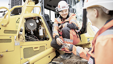 Zwei STRABAG Lernende in orangefarbener Schutzkleidung und Helm bei der Wartung eines gelben Baumaschinengeräts. Ein:e Lernende:r übergibt ein Werkzeug an den:die Kolleg:in, der:die in der Maschinenöffnung kniet. Im Hintergrund ist der Innenraum der Maschine sichtbar. 