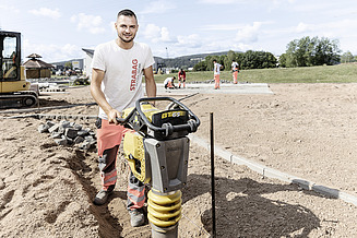 Lehre bei STRABAG - Schnuppere selbst rein Ein STRABAG Lernender in weissem T-Shirt und orange-grauer Arbeitshose bedient einen gelben Vibrationsstampfer auf einer Baustelle. Im Hintergrund sind weitere Mitarbeitende beim Verlegen von Pflastersteinen, ein Bagger sowie grüne Wiesen und ein Pavillon sichtbar.