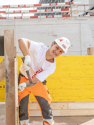 Junger STRABAG Lernender (Maurer) auf einer Baustelle. Er lächelt in die Kamera und hält einen Hammer und Nagel in der Hand. Mit weissem Helm und Shirt mit STRABAG Logo. Im Hintergrund sieht man eine Hochbaubaustelle