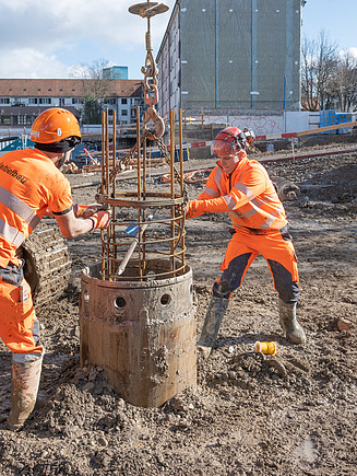 Ein STRABAG Lernender und ein STRABAG Polier in oranger Schutzkleidung bei der Baustellenarbeit. Sie befestigen eine Stahlbewehrung, die mit einem Kran angehoben wird. Im Hintergrund sind Gebäude und Baustellenmaterialien sichtbar. 