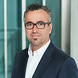 STRABAG Ausbildungskoordinator Fläche Schweiz STRAGAB Ausbildungskoordinator schaut lächelnd in die Kamera. Er ist mit Sakko und Hemd gekleidet und trägt eine Brille. Im Hintergrund ist ein Lift zusehen.