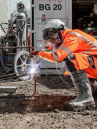 Ein STRABAG Lernender in oranger Schutzkleidung beim Schweissen auf einer Baustelle. Er trägt einen Schutzhelm mit Visier und bearbeitet eine Bewehrung, während Funken sprühen. Im Hintergrund ist noch mehr Baustellenequipment zu sehen.