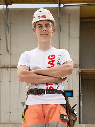 STRABAG Lernender in weissem STRABAG-T-Shirt, Helm und orangefarbener Arbeitshose steht selbstbewusst mit verschränkten Armen auf einer Baustelle. Er trägt einen Werkzeuggürtel mit verschiedenen Werkzeugen. Im Hintergrund sind ein Rohbau mit Beton- und Ziegelwänden sowie Gerüste zu sehen. 