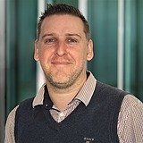STRAGAB Ausbildungsbeauftragter schaut lächelnd in die Kamera. Er ist mit einer Weste und Hemd gekleidet. Im Hintergrund ist ein Lift zusehen.