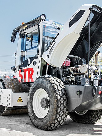 Zwei STRABAG-Mitarbeiter inspizieren einen weisse Walzenzug. Die Motorhaube ist geöffnet und zeigt den Innenraum der Maschine. Im Hintergrund sind Werkstatthallen und Maschinen zu sehen.