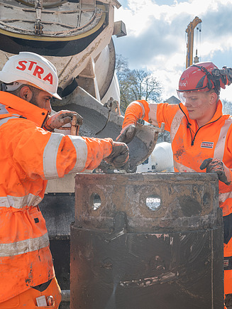 STRABAG Lernender mit STRABAG Polier in leuchtend oranger Schutzkleidung und Helmen bei der Arbeit an einer grossen Baumaschine – umgeben von Schlamm und Baustellengeräten, konzentriert im Austausch.