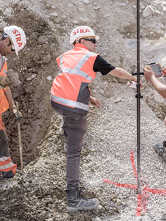 Drei STRABAG-Mitarbeitende in Schutzkleidung und Helmen bei der Vermessungsarbeit auf einer Baustelle. Einer führt Vermessungen durch, während die anderen mit Werkzeug und Messinstrumenten arbeiten. Im Hintergrund ist der Erdboden sichtbar, der für die Bauarbeiten vorbereitet wurde.