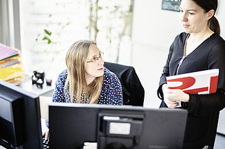 Zwei Mitarbeiterinnen in einem Büro im Gespräch. Die sitzende Person arbeitet an einem Computer und schaut die stehende Kollegin fragend an. Die stehende Person hält eine rote Mappe in der Hand und trägt schwarze Kleidung. Im Hintergrund sind Pflanzen und Fenster mit Blick nach draussen sichtbar. 
