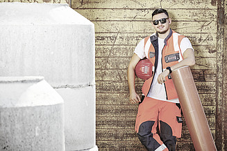 STRABAG Lernender in orangener Arbeitskleidung lehnt entspannt an einer rostigen Metallwand. Er trägt eine Sonnenbrille und hält einen roten Bauhelm unter dem Arm, während er sich auf ein grosses Kunststoffrohr stützt. Im Vordergrund sind Betonbauteile sichtbar. 