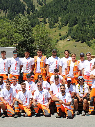 Eine grosse Gruppe von STRABAG-Lernenden, posiert gemeinsam in Arbeitskleidung und weissen STRABAG-T-Shirts vor einer idyllischen Berglandschaft. Einige tragen orange Sicherheitswesten, andere Sonnenbrillen und Kappen. Im Hintergrund sind grüne Hügel, Wälder und ein weites Tal zu sehen.
