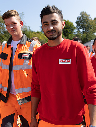 Eine Gruppe junger STRABAG-Lernender steht draussen in Arbeitskleidung beisammen und lächelt in die Kamera.