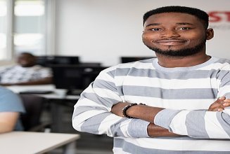 Kaufmännischer Lehrling mit lächelndem Blick in die Kamera und verschränkten Armen. Im Hintergrund arbeiten weitere Lernende an ihrem PC’s