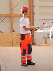Junger STRABAG Lernender (Zimmermann) beim Bewegen von Holzbalken mit Hilfe eines Krans, welchen er mit einer Fernbedienung steuert. Er trägt ein Helm zur Sicherheit. Im Hintergrund kann man die Holzbau-Werkstatt sehen.