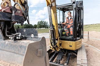 STRABAG Lernende in orangener Arbeitskleidung sitzt lächelnd in der Kabine eines Baggers auf einer Baustelle. Sie hält den Steuerhebel fest in der Hand, während die Baggerschaufel im Vordergrund sichtbar ist. Im Hintergrund erstreckt sich eine offene Landschaft mit Bäumen und einem klaren Himmel. 