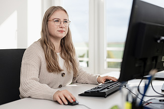 Eine STRABAG Lernende mit langen, blonden Haaren sitzt lächelnd an einem Schreibtisch in einem modernen Büro. Sie telefoniert und macht gleichzeitig Notizen. Auf dem Schreibtisch befinden sich ein Computer, eine Tastatur, ein Telefon sowie eine Tasse mit STRABAG-Logo. 