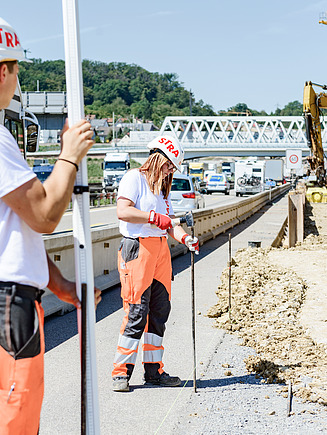 Zwei STRABAG Lernende bei Vermessungsarbeiten an einer Autobahnbaustelle. Einer hält eine Vermessungsstange, während die andere Markierungen setzt. Im Hintergrund sind Verkehr, Brücken und Baustellenfahrzeuge sichtbar.