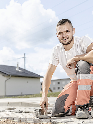 Ein STRABAG Lernender in Arbeitskleidung kniet auf einer Baustelle und setzt Pflastersteine. Er trägt ein weisses T-Shirt und orange-graue Arbeitshosen. Im Hintergrund sind ein Wohnhaus, ein Pavillon sowie Strommasten zu sehen.