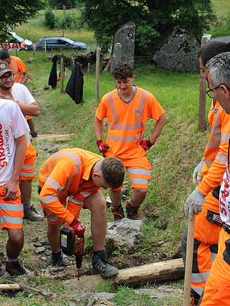 Eine Gruppe von STRABAG Lernenden in orangener Arbeitskleidung arbeitet gemeinsam an einem Hang. Einer Lernender benutzt eine Bohrmaschine, während die anderen ihn unterstützen und das Gelände vorbereiten. Im Hintergrund sind grüne Wiesen und Bäume.