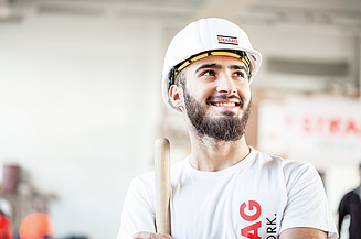 Schnupperlehre bei STRABAG STRABAG Lernender mit weissem Bauhelm und einem weissen STRABAG T-Shirt steht lächelnd auf einer Baustelle. Er hält ein Werkzeug in der Hand und blickt motiviert in die Ferne. Im Hintergrund sind unscharf Baustellenstrukturen und ein STRABAG-Banner zu erkennen.