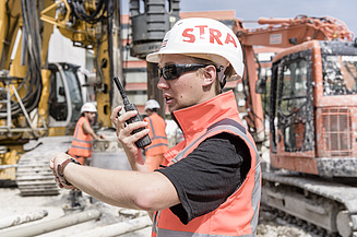 Auszubildender Grundbauer mit Funkgerät in der Hand auf der Baustelle mit Baumaschinen im Hintergrund