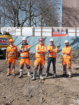Fünf STRABAG-Mitarbeiter in oranger Schutzkleidung stehen selbstbewusst auf einer Baustelle. Die Gruppe posiert vor einem Bagger und Bauabsperrungen, im Hintergrund sind Bauzaunbanner und Baustellenzäune sichtbar.