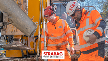 STRABAG Lernender mit weiterem Bauarbeiter in leuchtend oranger Schutzkleidung und Helmen bei der Arbeit an einer grossen Baumaschine – umgeben von Schlamm und Baustellengeräten, konzentriert im Austausch.