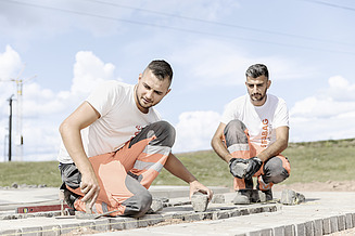 Schnupperlehre im Strassenbau Zwei STRABAG Lernende in weissen T-Shirts und orange-grauen Arbeitshosen setzen konzentriert Pflastersteine auf einer Baustelle. Sie knien auf dem Boden und platzieren die Steine präzise in die vorbereitete Fläche. Im Hintergrund sind blauer Himmel, Wolken und ein Kran sichtbar.