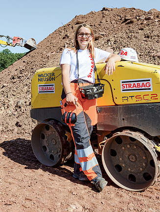 Drei Lernende von STRABAG auf einer Baustelle – zwei im Gespräch, eine Person lehnt an einer Bodenwalze und lächelt in die Kamera.