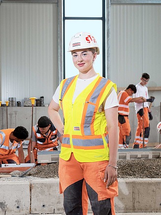 Lernende von STRABAG arbeiten in einer Halle an einer Baustelle mit Kies – im Vordergrund steht eine junge Frau mit Helm und Warnweste und blickt selbstbewusst in die Kamera.