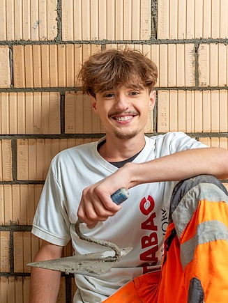 Maurerlehrling sitzt lächelnd in die Kameran blickend mit angewinkelten Knien vor einer Lochsteinmauer mit einer Maurerkelle in der Hand.