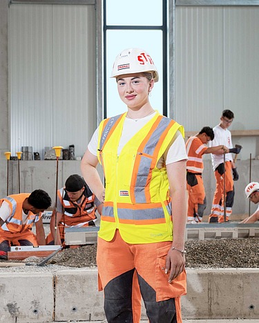 Eine Gruppe von STRABAG-Mitarbeitenden, darunter Auszubildende und Fachkräfte, in Arbeitskleidung. Im Vordergrund stehen junge Lernende in roten und orangefarbenen Sicherheitswesten. Im Hintergrund sind Baugeräte sowie grüne Bäume sichtbar. Die Stimmung wirkt motiviert und teamorientiert.