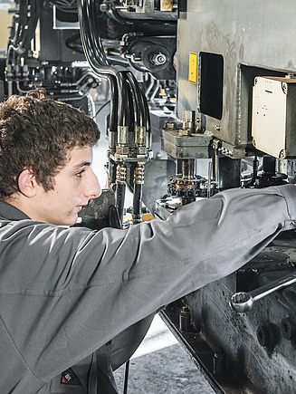 Ein STRABAG Lernender richtet Industriemaschine ein. Er trägt graue Arbeitskleidung und konzentriert sich auf die Einstellung der Teile.