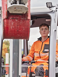 Junger STRABAG Lernender in einem Bagger bei der Arbeit. Er trägt orange-leuchtende Arbeitskleidung.