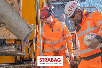 STRABAG Lernender mit weiterem Bauarbeiter in leuchtend oranger Schutzkleidung und Helmen bei der Arbeit an einer grossen Baumaschine – umgeben von Schlamm und Baustellengeräten, konzentriert im Austausch.